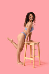 Wall Mural - Beautiful young happy African-American woman in stylish underwear with chair on pink background