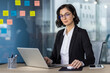 © Liubomir - Confident businesswoman in glasses working on laptop in modern office. Colorful sticky notes on glass board in background. Concept of productivity, determination, and professional work environment.