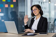 © Liubomir - Smiling woman in formal business attire participating in video conference from modern office setting. Businesswoman engaging in virtual meeting, waving at laptop, demonstrating communication