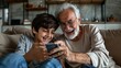 © Drew - An elderly man and his grandson are engrossed with a smartphone, sharing a moment of laughter and joy on a comfortable sofa. The scene highlights their special bond.