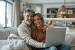 © btiger - Happy middle aged couple on sofa using laptop relaxed at home smiling laughing mature man and woman in sunny living room having fun candid shot