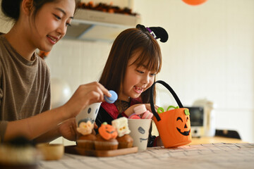 Cheerful little girl and mother decorating Halloween cupcakes with different monsters at kitchen table