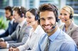 © Sirinporn - Smiling help desk representative sitting at a desk with a headset on, surrounded by cheerful coworkers blurred in the background, exemplifying excellent teamwork and customer support.