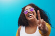 © Sanc/peopleimages.com - Portrait, happy and woman with peace sign in studio for gen z fashion, heart glasses and aesthetic style. Excited, girl and cool v icon with trendy eyewear, emoji and sunglasses on blue background