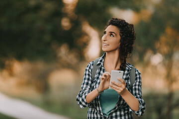 woman with curly braided hair and a checkered shirt smiles while holding a smartphone, looking up. Her casual attire and backpack suggest she is enjoying a moment of outdoor photography