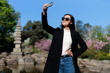 Wall Mural - A woman is taking a selfie with her cell phone in a park. She is wearing a black coat and blue jeans