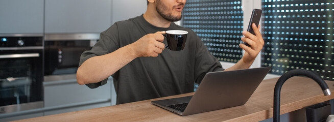 Wall Mural - Panoramic portrait of young satisfied  man working home at laptop on kitchen, looking in smartphone with, holding coffee cup. Business concept. Wearing eyeglasses and sweater.