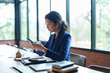 © snowing12 - A house seller in a blue suit is focused on her phone while sitting at a desk. The bright, natural light from the large windows illuminates her workspace.