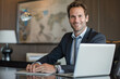 © forenna - Professional portrait of a successful male businessman seated at his desk, using a laptop and smiling confidently at the camera, perfect for illustrating financial expertise and co