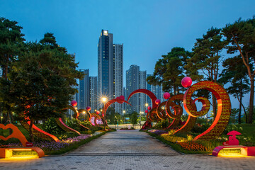 Seo-gu, Incheon, South Korea - July 6, 2023: Night view of red light on heart shape sculptures on sidewalk blocks with pine trees against apartments at Cheongna Lake Park
