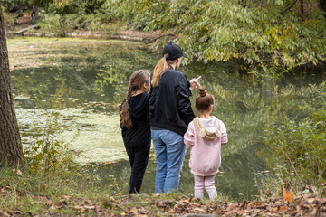 A heartwarming scene of a Caucasian mother and her two young daughters, one with blonde hair and the other with brown hair, enjoying a peaceful moment by a serene lake in a lush, green forest.