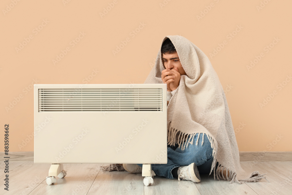 Frozen young man with plaid and radiator sitting near beige wall