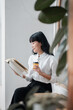 © Satori Studio - A young woman in a white shirt reads a book and drinks coffee in a modern, minimalist interior with natural light.
