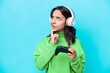 © luismolinero - Young hispanic woman playing with a video game controller isolated on blue background having doubts while looking up