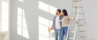 © Studio Romantic - Family couple doing renovations at home. Young man and woman change color of walls in living room. Banner with happy husband and wife standing by ladder and looking at freshly painted light gray wall