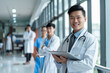 © GustavsMD - Handsome Asian male doctor with a stethoscope, holding a tablet and smiling confidently in a hospital corridor with medical staff in the background