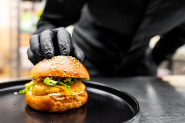 Wall Mural - Close-up of a fresh burger with a sesame bun and greens on a black plate, held by a person in black attire with gloves, against a blurred background