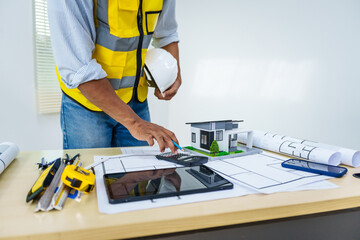 Asian male architect works at desk with house model toy and A3 paper blueprints, construction real estate law, zoning, permits, and resolving property disputes through arbitration and litigation.