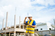 © NanSan - An architect, wearing a hardhat and safety vest, checks a laptop and house plan paper on a clipboard. Blueprints and construction documents lie nearby, emphasizing safety and precision in building.