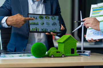 Two Asian businessmen in formal suits discuss ideas and suggestions at an office desk, focusing on green housing estates, city planning, renewable energy, and achieving net zero emissions by 2050.