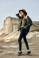 Wall Mural - Stylish young woman in plaid shirt and jeans standing confidently in the desert wearing a hat