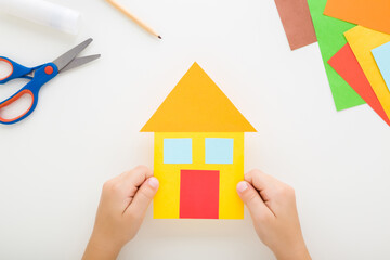 Wall Mural - Little child hands holding house shape from colorful application paper on light gray table background. Making family home. Point of view shot. Closeup. Top down view.