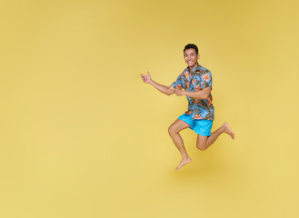 Wall Mural - Full body young asian tourist man wear beach shirt jumping and pointing finger isolated on yellow copy space background studio. Summer vacation sea rest sun tan concept