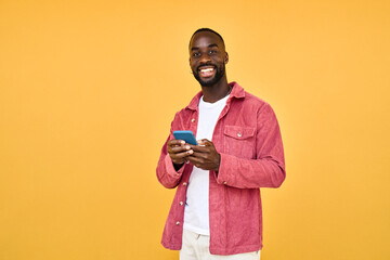 Young happy smiling African American Black hipster man wearing red shirt with cellphone standing isolated on yellow background holding smartphone using mobile cell phone technology looking at camera.