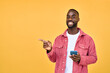 © insta_photos - Young happy African American Black hipster man with cellphone in hands standing isolated on yellow background holding smartphone using mobile cell phone pointing aside advertising betting app.