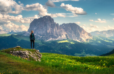 Wall Mural - Girl on the hill with yellow flowers and green grass in beautiful alpine mountain valley at sunset in summer. Landscape with young woman in alps, sky with clouds, rocks. Travel in Dolomites, Italy