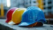 © Damerfie - A close-up view of three colorful construction helmets, in red, yellow, and blue, placed on a wooden surface at a construction site, representing safety and site readiness.