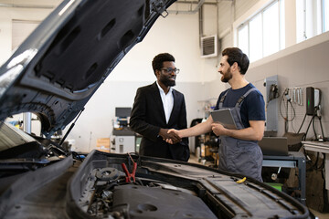 Mechanic in uniform shaking hands with customer in suit near car engine in auto shop. Concept of service, professionalism, satisfaction, and business relationship in automotive industry.