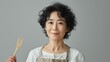 © RaptorWoman - Middle-aged woman in apron holding wooden fork, smiling and posing against a gray background.