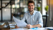 © Jack - Portrait of a young businessman smiling at the camera, sitting at a desk in the office and working with documents and an invoice