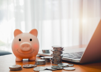 Online banking, saving account, profit, income, financial investment concept. Happy smile face of cute pink fat piggy bank and many coins piles near laptop computer on wooden table with copy space.