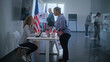 © Framestock - African American woman comes to registration table at polling station. Female polling worker talks to woman and uses digital tablet. Multiethnic US citizens vote in voting booths. Election Day in USA.