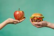© Maryam - A vivid comparison of unhealthy and healthy food choices; a hamburger held in one hand and a fresh apple in the other against a green background