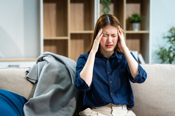 A young Asian woman sits on a sofa in her living room, feeling sick with a headache. She experiences throbbing pain and nausea, seeking comfort and relief at home.