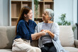 © Phushutter - A mature mom and a young Asian woman, mother and daughter, sit together on a sofa in their living room, celebrating Mother's Day with love and bonding at home.