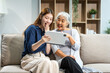 © Phushutter - A mature mom and a young Asian woman, mother and daughter, sit together on a sofa in their living room, celebrating Mother's Day with love and bonding at home.