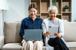 © Phushutter - In their living room, a young Asian woman and her grey-haired mature mom, mother and daughter, sit together on a sofa. daughter works online with laptop while mom lovingly cares and supports her.