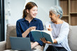 © Phushutter - In their living room, a young Asian woman and her grey-haired mature mom, mother and daughter, sit together on a sofa. daughter works online with laptop while mom lovingly cares and supports her.