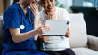 © Phushutter - A young Asian nurse cares for an older grey-haired woman on a sofa, assessing health and discussing osteoporosis, arthritis, Alzheimer's, and dementia, using a tablet for information.