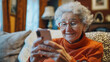 © thanakrit - Senior woman using a smartphone to video call family, smiling and sitting in a cozy living room.