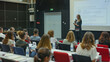 © VK Studio - A woman delivers a presentation to a seated audience, accompanied by a projector screen showcasing information in a brightly lit lecture room.