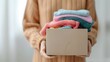 © May Chanikran - Person holding a cardboard box filled with folded colorful clothes. Concept of donation, sustainable living, and wardrobe organization.