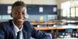 © Vadym - African American teen boy wears classy school uniform with blue jacket, black tie. Stands in front of desk with two chairs in gray classroom. Student smiles at camera for back to school theme.