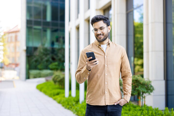 hispanic man in casual attire standing outside modern office building using smartphone. daylight, ur