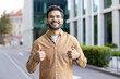 © Liubomir - Smiling hispanic man wearing casual outfit giving thumbs up on city street. Young professional showing positivity, confidence, and approval outdoors urban environment. Modern buildings background.
