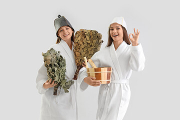 Canvas Print - Young women in sauna hats with whisks and bucket of water on light background